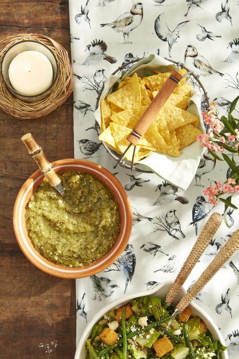 a bowl of roasted tomatillo and poblano salsa with a small spoon and a wire basket full of tortilla chips next to it