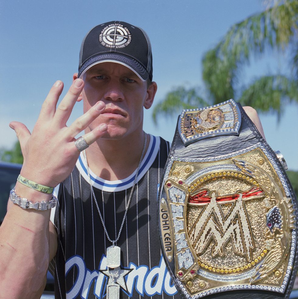 john cena holds a bejeweled wrestling belt up with one hand and flashes his other palm near his face, he wears a black ball cap and an orlando jersey