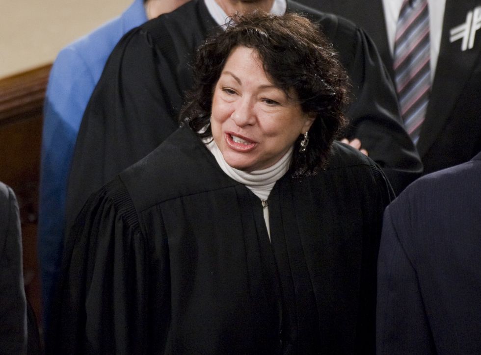 sonia sotomayor, wearing black judge robes, speaking to someone off camera in the middle of a crowded room