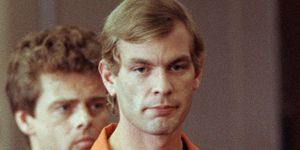 jeffrey dahmer wears an orange prison uniform shirt with a black undershirt, he has a neutral expression on his face, behind him on the left is a police officer