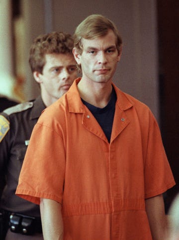 jeffrey dahmer wears an orange prison uniform shirt with a black undershirt, he has a neutral expression on his face, behind him on the left is a police officer