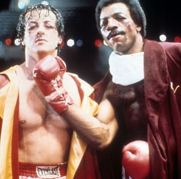 carl weathers pretending to give sylvester stallone an uppercut punch for the movie rocky, both men wear robes around their shirtless bodies, weathers has on red boxing gloves
