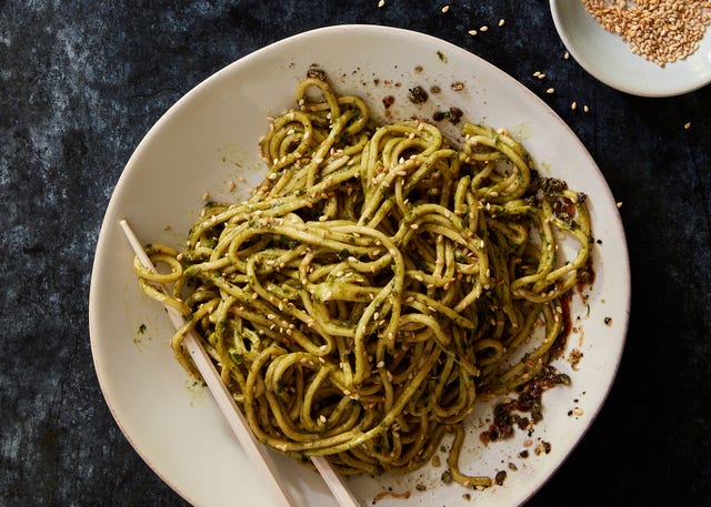 a white bowl of noodles in green pesto sauce topped with toasted sesame seeds
