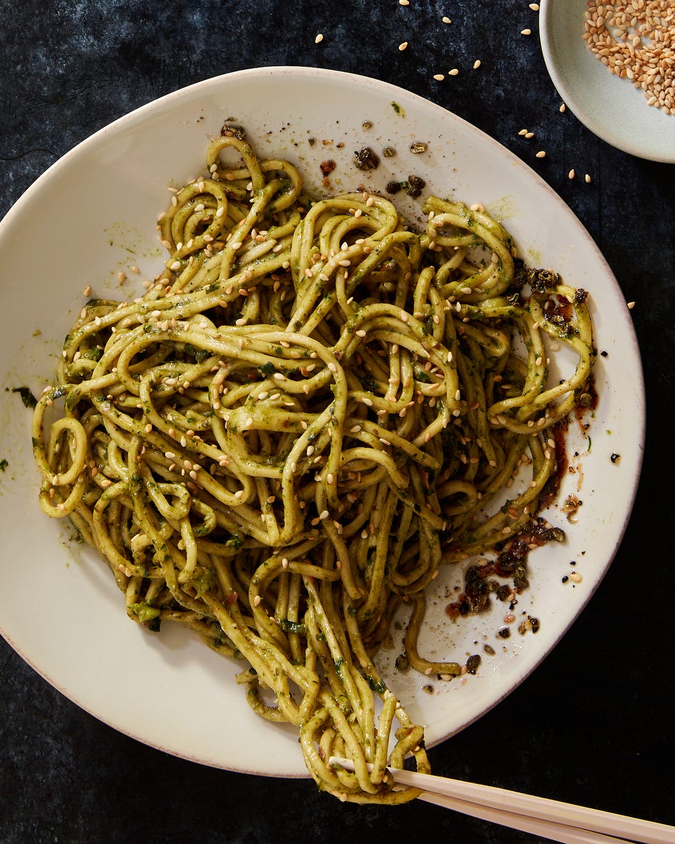a white bowl of noodles in green pesto sauce topped with toasted sesame seeds