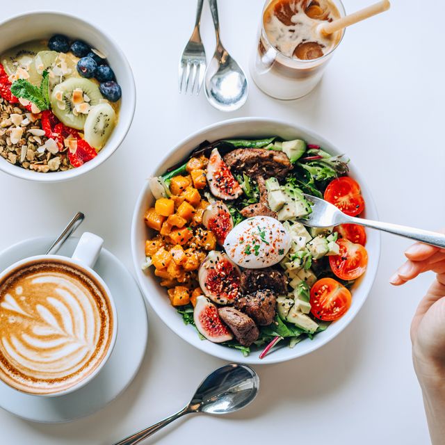 table top view of a woman eating healthy beef cobb salad bowl with greens, avocado, cherry tomatoes, figs, pumpkin and soft boiled egg, vegan chia and quinoa granola fruit bowl and coffee by the side healthy food trend nutritious and well balanced diet