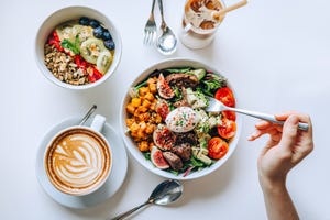 table top view of a woman eating healthy beef cobb salad bowl with greens, avocado, cherry tomatoes, figs, pumpkin and soft boiled egg, vegan chia and quinoa granola fruit bowl and coffee by the side healthy food trend nutritious and well balanced diet