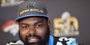 michael oher looks at the camera while sitting in front of a microphone, he wears a carolina panthers super bowl 50 hat and his panthers jersey