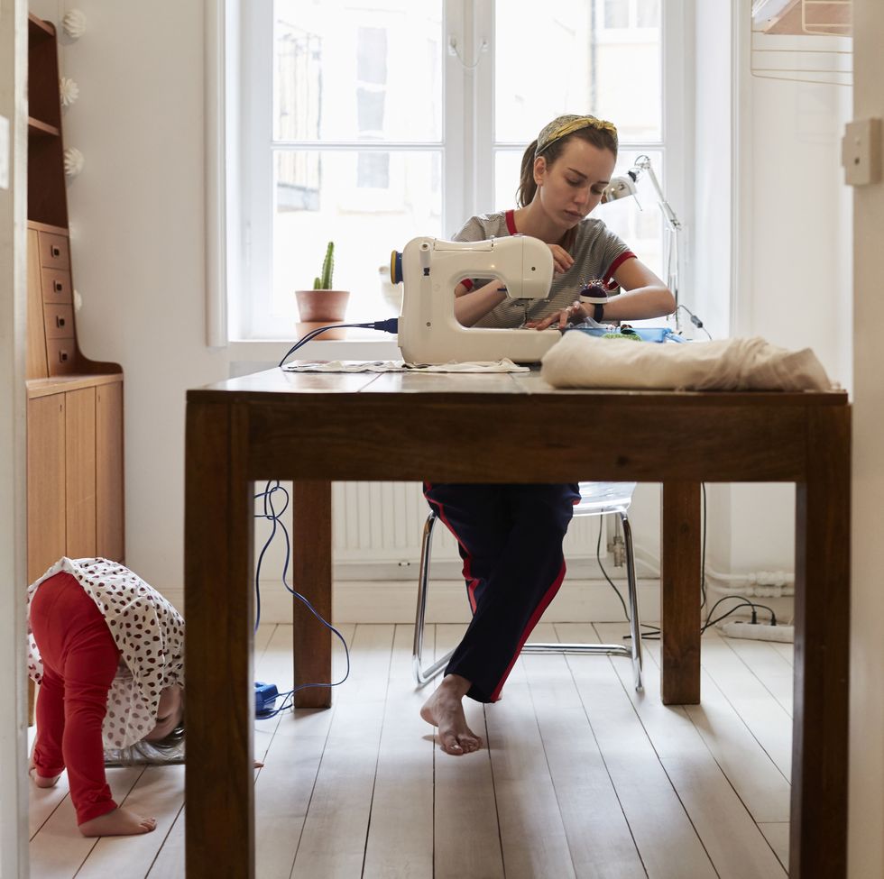 jobs for stay at home moms - Tailor sewing at table while daughter playing on floor seen through doorway at home