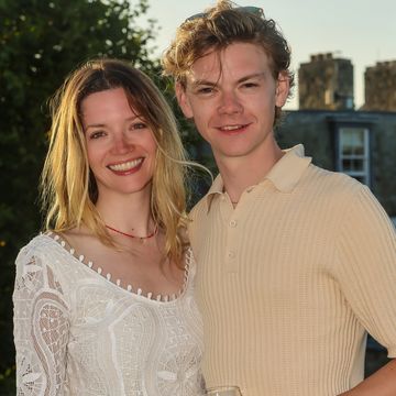 talulah riley and thomas brodie sangster, a young man and woman stand hugging as they smile at the camera, she has blonde hair worn down and wears a white dress, he has light brown curly hair and wears a beige top and trousers