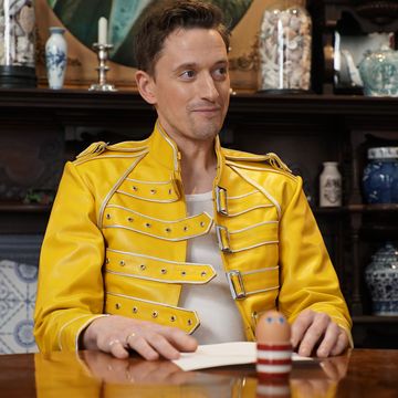 john robins sits inside the taskmaster house's task room, in front of a table containing a task and an egg in a striped cup with googly eye stickers on it