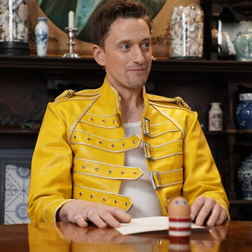 john robins sits inside the taskmaster house's task room, in front of a table containing a task and an egg in a striped cup with googly eye stickers on it