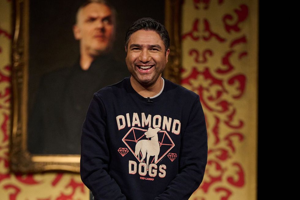 nick mohammed smiles as he stands on the taskmaster stage in front of a small pushchair containing a soft toy doll custom made in his likeness