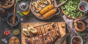 tasty grilled bbq meat ribs on wooden cutting board and grill grate with knife and fresh garlic herb butter bread served on rustic table with grilled corn cobs with bbq sauce , salad leaves and other tasty ingredients