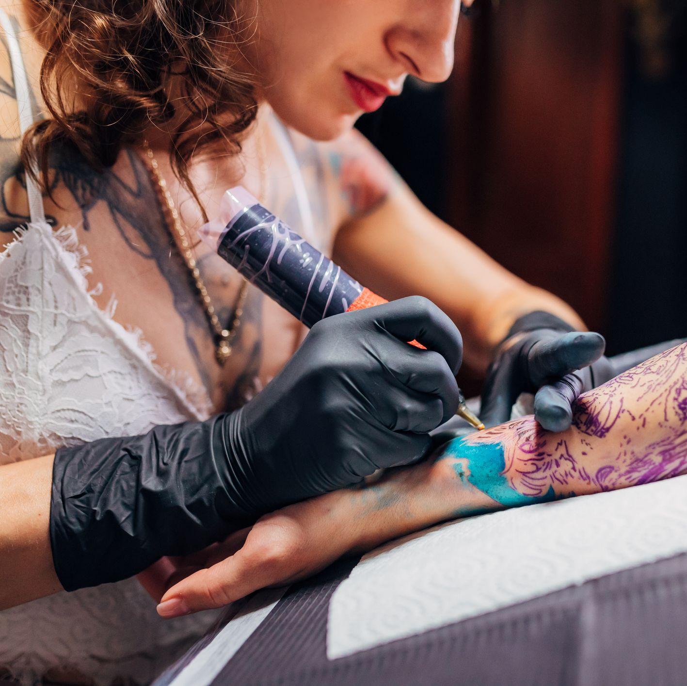 tattoo artist wearing black latex gloves tattooing colourful tattoo on woman's visible outstretched arm