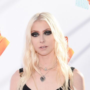 taylor momsen, a young woman stands looking at the camera, she has long blonde hair worn down in loose waves, heavy black eye makeup and wears a short black dress