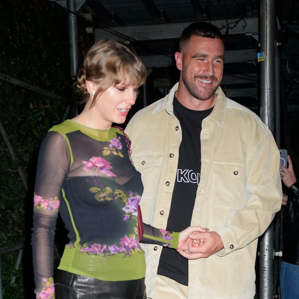 taylor swift and travis kelce walk hand in hand on a city sidewalk, she wears a sheer floral long sleeve top with a black skirt, he wears a tan jacket over a black t shirt and matching tan pants