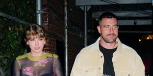 taylor swift and travis kelce walk hand in hand on a city sidewalk, she wears a sheer floral top with a black mini skirt and boots, he wears matching tan corduroy pants and a top over a black shirt