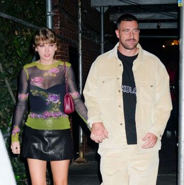 taylor swift and travis kelce walk hand in hand on a city sidewalk, she wears a sheer floral top with a black mini skirt and boots, he wears matching tan corduroy pants and a top over a black shirt