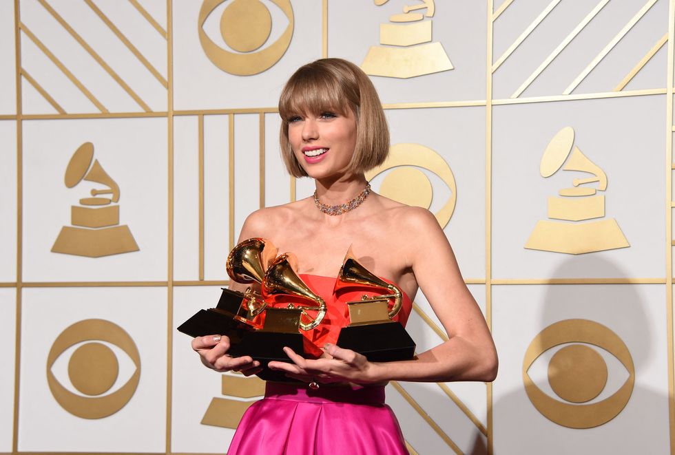 taylor swift smiles and stands while holding three grammy awards, she wears a strapless orange and hot pink gown with a gold chocker necklace
