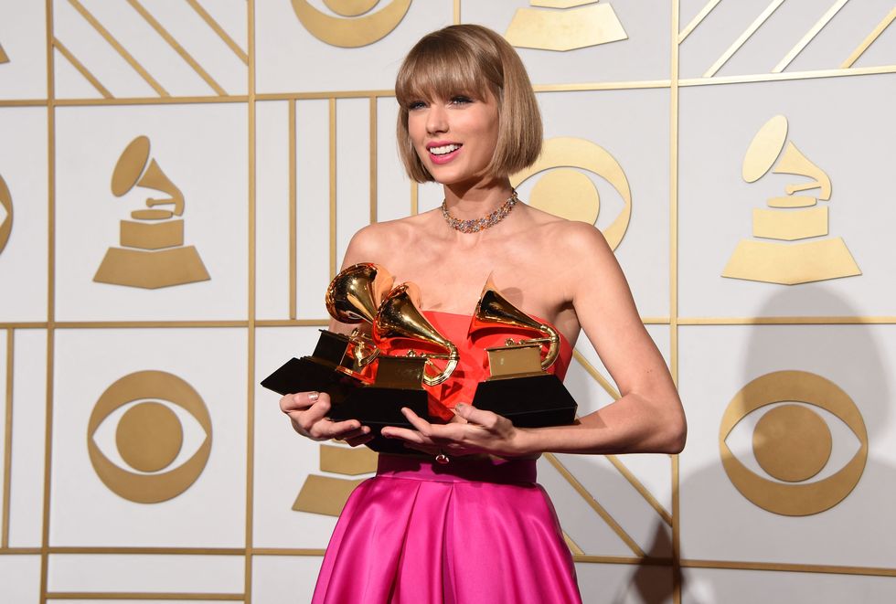 taylor swift smiles and stands while holding three grammy awards, she wears a strapless orange and hot pink gown with a gold chocker necklace