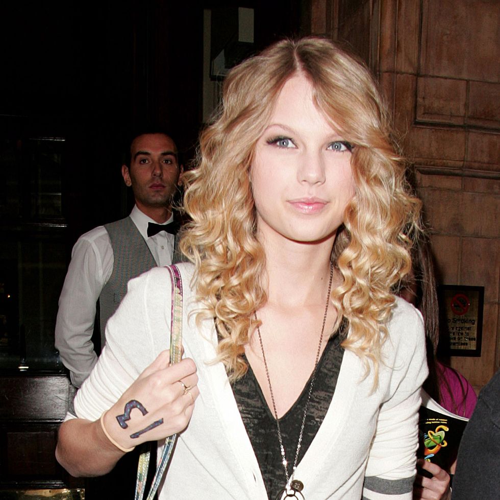 a woman with curly hair smiles as she walks and holds the strap of a bag on her shoulder, her hand has the number 13 written on it