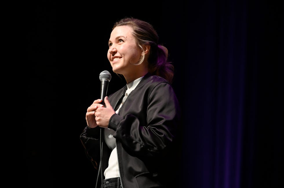 taylor tomlinson smiles while looking to the left and holding a microphone in front of her face, she wears a black jacket over a white shirt and black pants