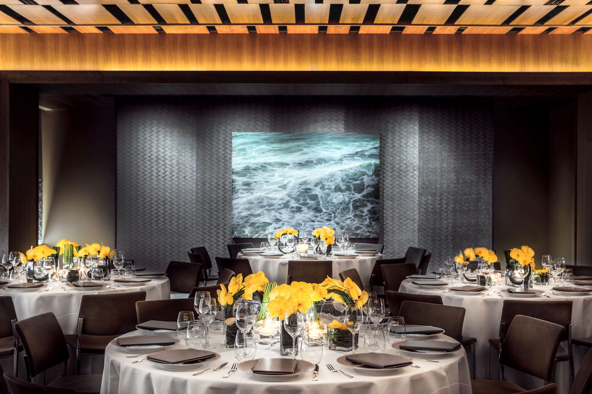 fully set round tables with yellow orchids in a dining room with a painting of the ocean at prive by le bernardin, nyc