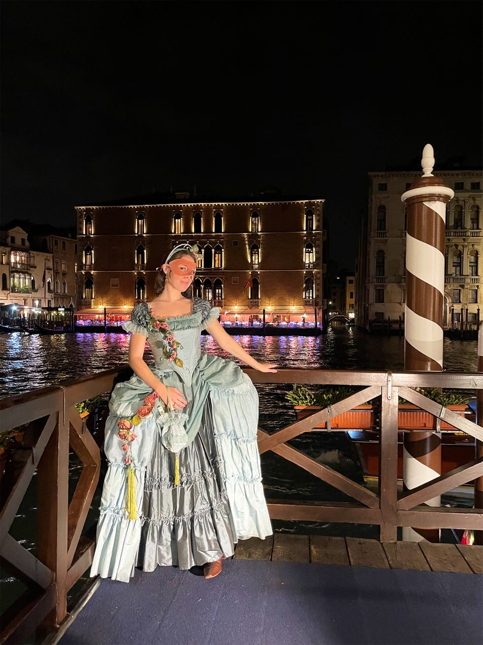 person in formal attire posing near a canal at night with a historical building in the background