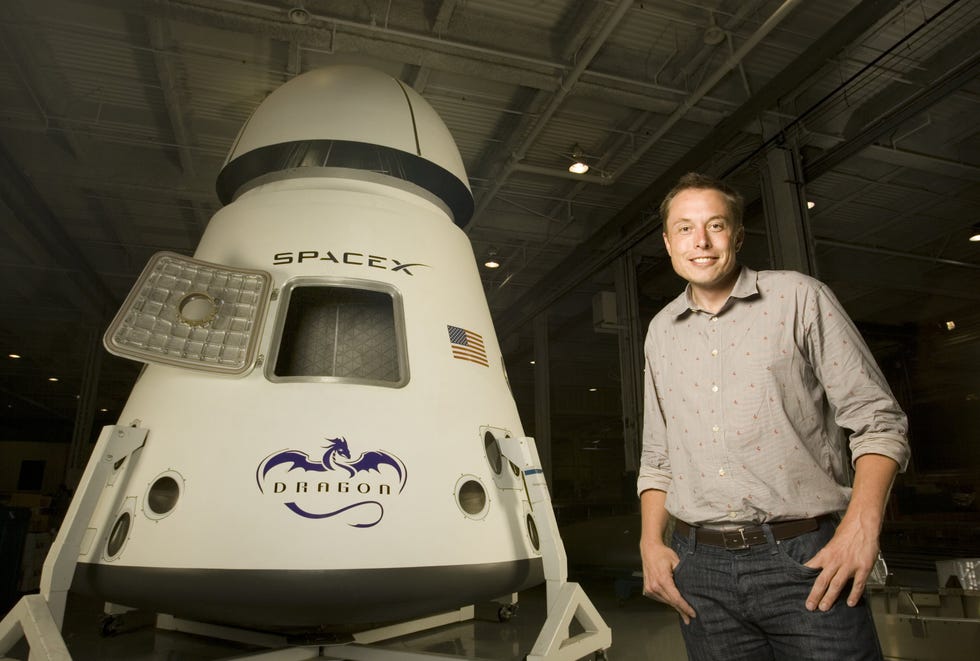 elon musk poses for a portrait next to a spacecraft, he wears a collared button up shirt and jeans with a belt