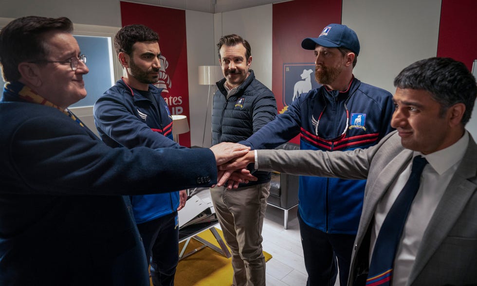 In einer Szene aus Ted Lasso versammeln sich Higgins, Roy, Ted, Beard und Nate im Trainerzimmer zu einem Ritual vor dem Spiel