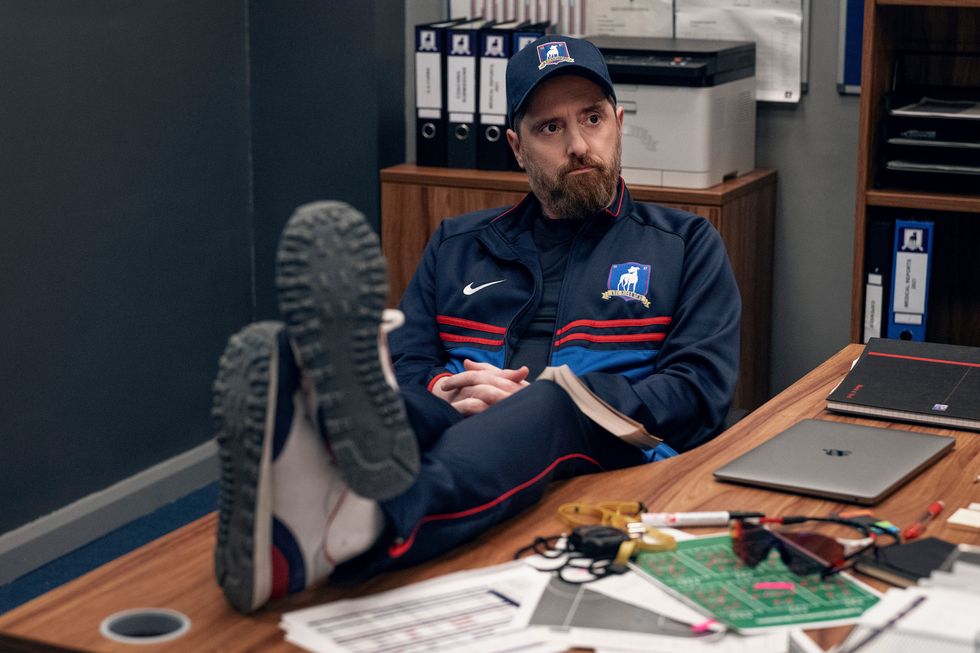 en una escena de la temporada 3 de ted lasso, el entrenador barba se sienta con los pies sobre el escritorio en la oficina del equipo