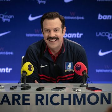 a publicity still of jason sudeikis portraying ted lasso, wearing a blue athletic jacket, sitting at a table with two microphones and several cellular phones and recording devices, with the words we are richmond in the front, and a blue backdrop with various advertising logos in the background