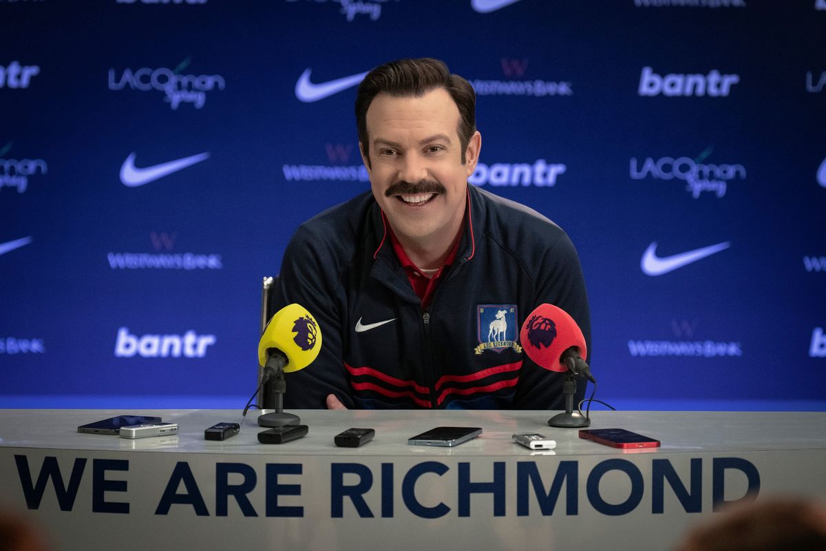 a publicity still of jason sudeikis portraying ted lasso, wearing a blue athletic jacket, sitting at a table with two microphones and several cellular phones and recording devices, with the words we are richmond in the front, and a blue backdrop with various advertising logos in the background