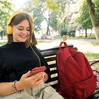 teen with yellow headphones sitting on bench with red backpack