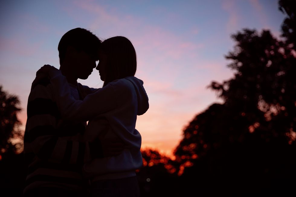 teenage couple in silhouette against a pink and orange sky at sunset on a summer evening in a non urban scene