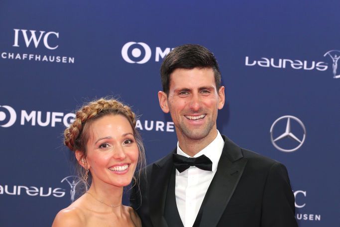 jelena and novak djokovic pose for a photo and smile at the camera, she wears a gray strapless stress and he wears a black tuxedo