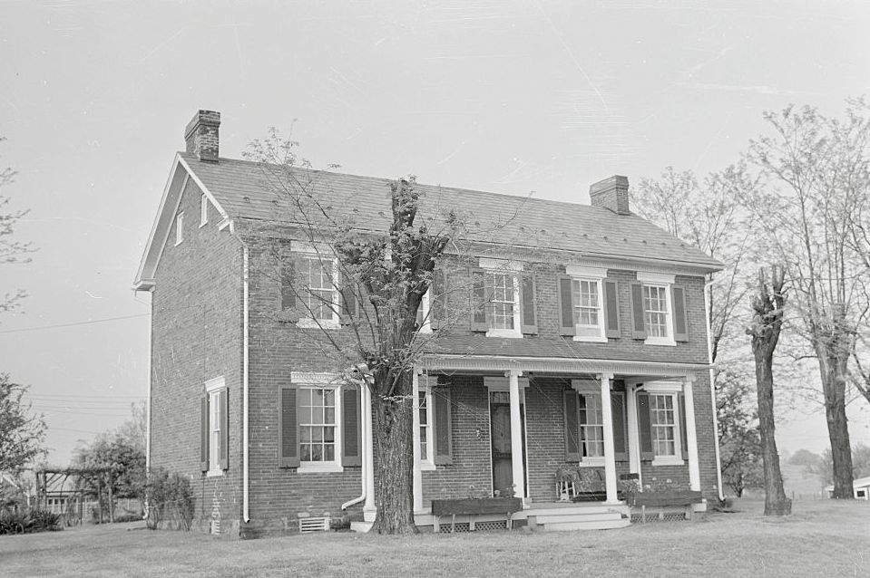 a brick house with a covered front porch sits in the foreground, it has a lawn with several trees surrounding it