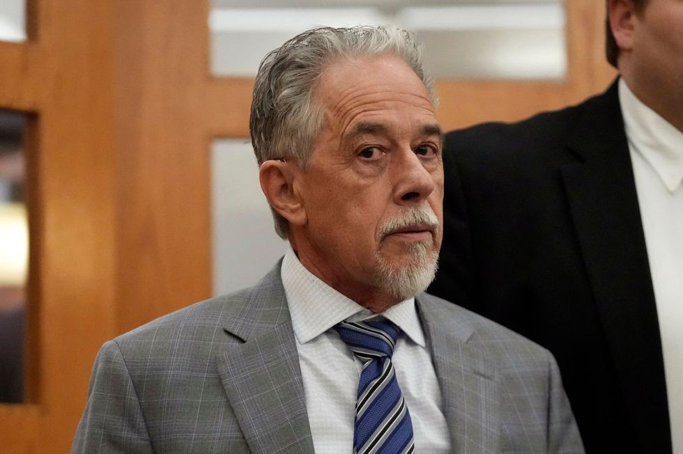 terry sanderson looks sideways at the camera with a solemn expression, he has graying hair and a white mustache and goatee, he is wearing a gray suit jacket with a subtle pattern, a white collared shirt with a subtle pattern and a blue tie with black and white stripes, behind him are parts of two wooden doors with frosted glass panes