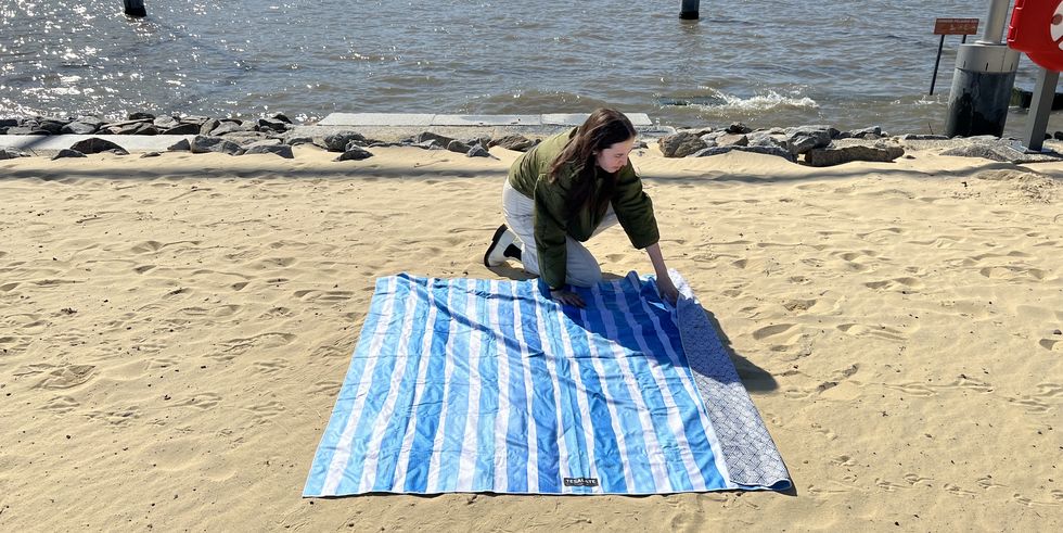 a good housekeeping analyst setting up a tesalate beach towel with blue and white stripes on the sand beach, good housekeeping's testing for the best beach towels