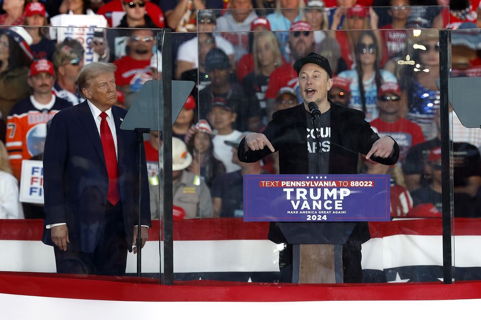 donald trump stands on a stage and watches elon musk talk at a podium, a crowd of people sits behind