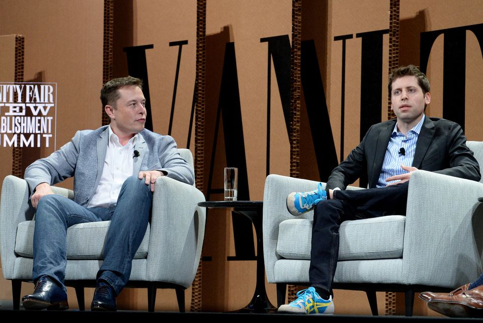 two men sitting in armchairs on a stage and talking, a side table rests in the middle of the chairs