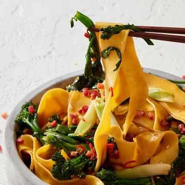a bowl of thick wide noodles, broccolini, scallions, pancetta, and red chilis being picked up by chopsticks