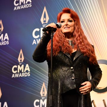 wynonna judd standing at a staged microphone and smiling for a photo