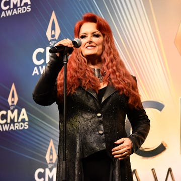 wynonna judd standing at a staged microphone and smiling for a photo