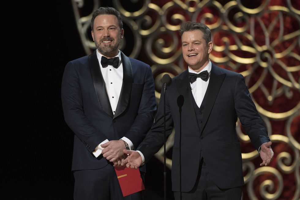 a bearded ben affleck, wearing a black tuxedo, holds a red envelope and stands next to matt damon, also wearing a black tuxedo, on a stage in front of a microphone