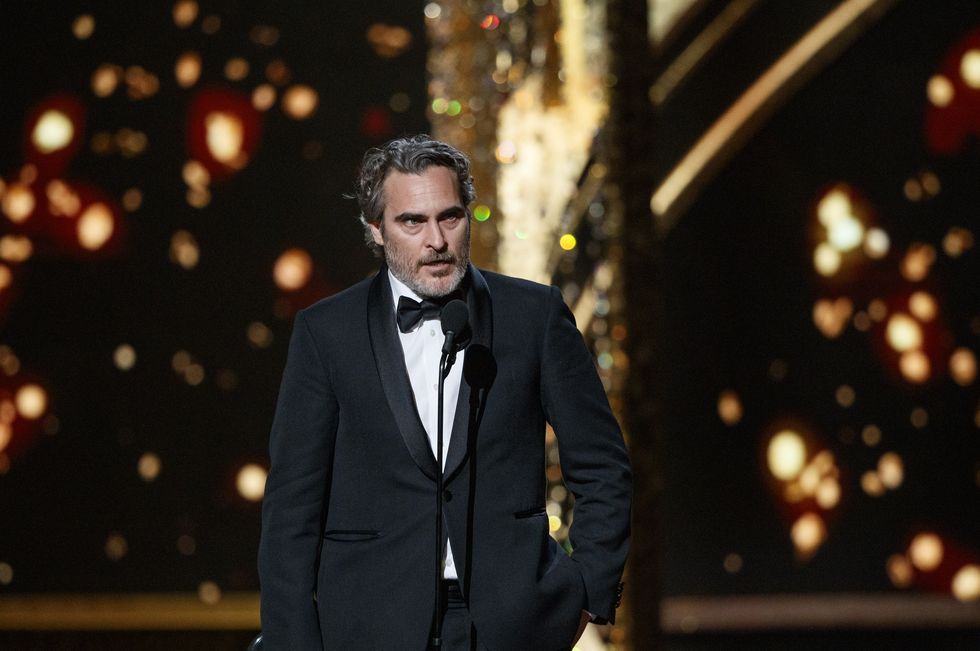 joaquin phoenix speaks into a microphone while standing on a stage, he wears a black suit with a black bowtie and white collared shirt, one hand holds an oscar statuette, his other hand is in his pocket