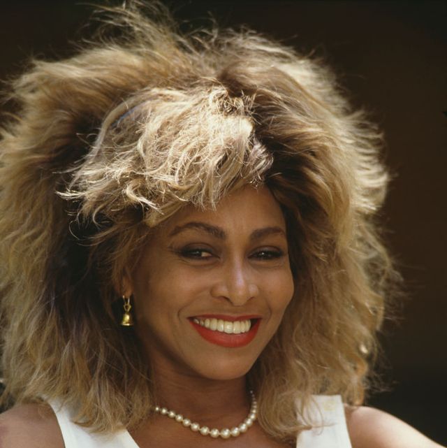 tina turner smiles at the camera, she wears a pearl necklace, gold bell earrings, a white sleeveless top, and red lipstick