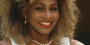 tina turner smiles at the camera, she wears a pearl necklace, gold bell earrings, a white sleeveless top, and red lipstick