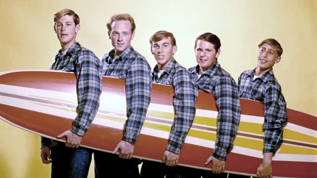 five young men wear matching plaid shirts and hold a red and yellow surfboard