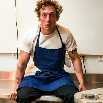 the bear, jeremy allen white sitting on top a metal work surface in a blue chef apron, looking stressed